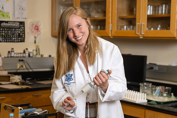 A white female student in a white lab coat looks towards the camera in a lab.