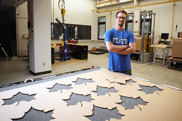 A white male student with a blue shirt standing in front of a large machine with a sheet of cardboard with Sycamore leaves cut out.