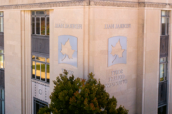 Aerial image of the corner of a large square building with a large metallic indiana state university sycamore leaf logo and the words “federal hall” above the leaf and “scott college of business” below the leaf with green trees in the foreground and on the side of the building.