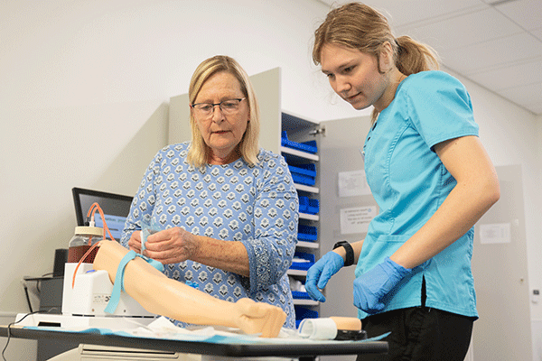  a White blond female student checks the blood pressure of a patient with back turned to the camera. 