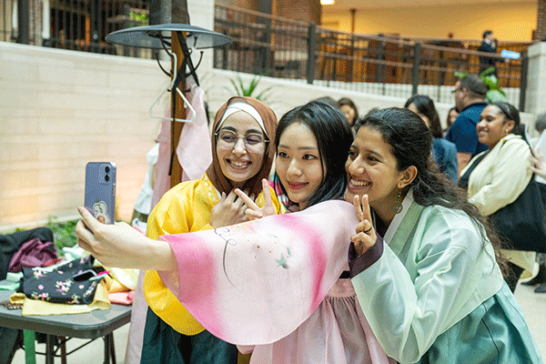 Three female students of diverse international heritage pose for a selfie during a cultural celebration event on campus. They wear colorful, billowing clothing of green, pink, and yellow hues. 